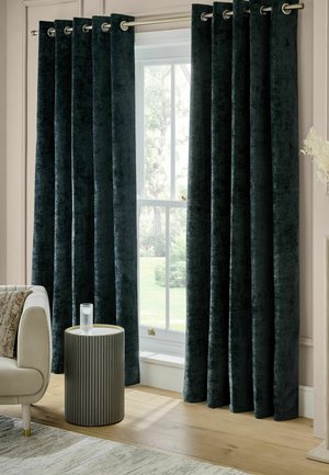 Dunkelblaue Samtvorhänge mit Metallösen hängen offen an einem Fenster neben einem beigen Sofa und einem runden Beistelltisch mit einem Glas Wasser.