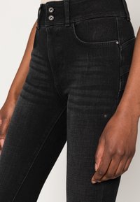 Pantalones vaqueros negros de mezclilla con ajuste slim, cintura alta, cierre de doble botón y bolsillos laterales. Presentan tela texturizada y detalles sutiles de costuras.