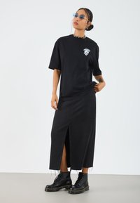T-shirt oversized noir avec graphique de crâne, associé à une jupe maxi en denim noir avec une fente sur le côté. Complété par des bottes montantes noires.