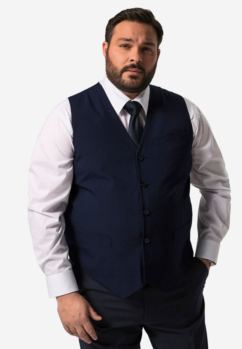 Gilet blu navy realizzato in tessuto dalla trama liscia, con chiusura a quattro bottoni, rever a punta e tasche laterali sopra una camicia bianca.
