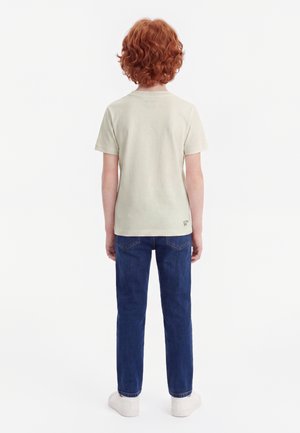 Niño con cabello rizado rojo está de espaldas, lleva una camiseta beige claro, vaqueros azules y zapatillas blancas sobre un fondo blanco.