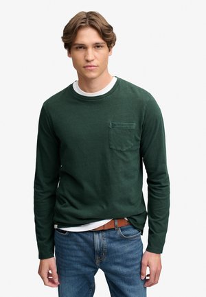 Ung mand med lyst brunt hår iført en mørkegrøn langærmet skjorte med brystlomme og blå jeans mod en ensfarvet hvid baggrund.