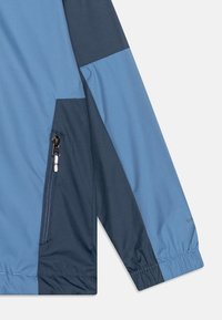 Giacca leggera con un mix di pannelli blu chiaro e scuro, tasca laterale con zip, polsini elastici e texture liscia.