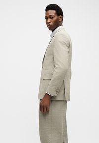 Άνδρας που φορά ανοιχτό μπεζ κοστούμι και άσπρο πουκάμισο, στέκεται πλευρικά, κοιτάζοντας ευθεία μπροστά με φόντο έναν απλό, ανοιχτόχρωμο τοίχο.