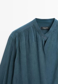 Blouse bleu sarcelle à coupe décontractée, dotée d'un col en V, de détails avec boutons et de plis froncés à l'épaule. Texture lisse et douce.