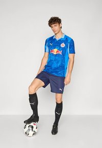 Kék focimez világoskék díszítésekkel és Red Bull logóval, sötétkék nadrággal, fekete zoknival és stoplival. A játékos labdával pózol.