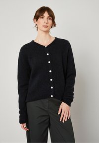 Tums zilas adītas kardigāns ar apaļu kakla izgriezumu un garām piedurknēm. Priekšpusē ir deviņas baltas pogas un maiga, strukturēta apdare.