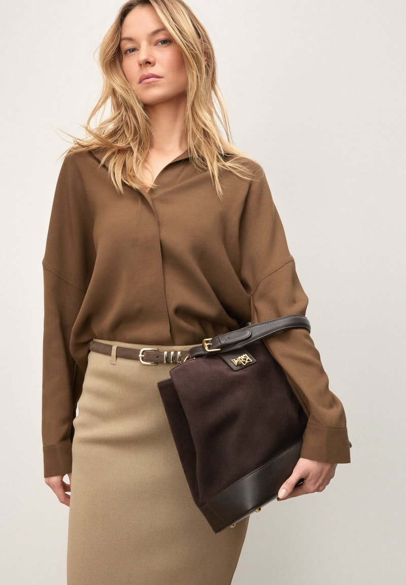 Femme en blouse marron et jupe beige tenant un grand sac à main marron foncé avec des détails en cuir sur un fond clair.