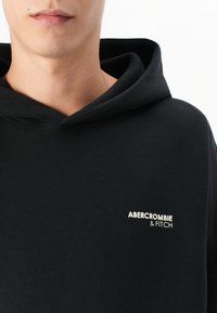 Sweat-shirt à capuche noire avec une texture douce, arborant un petit logo blanc "Abercrombie & Fitch" sur la poitrine. Design simple et coupe décontractée.