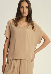Beige, loose-fitting top with a V-neck and short sleeves. Smooth texture, relaxed shape, and subtle draping. No prominent patterns or hardware.