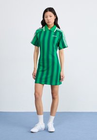Rochie polo verde cu dungi, cu guler, mâneci scurte și accente albe. Asortată cu adidași albi și șosete până la gleznă. Fundal simplu.