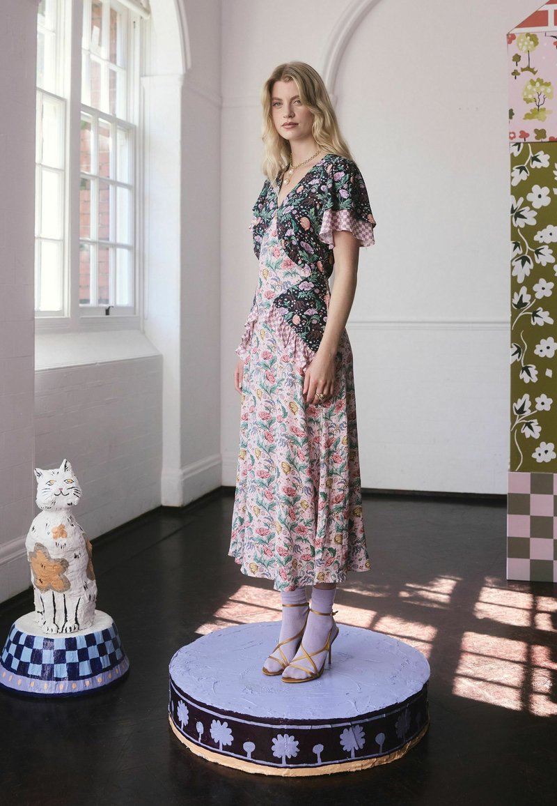 Žena oblečená v kvetinových patchwork šatách a levanduľových ponožkách s žltými podpätkami stojí na zdobenej okrúhlej platforme v jasnej miestnosti.