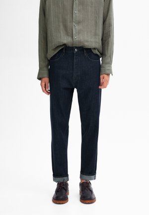 Person wearing cuffed dark blue jeans, olive green button-up shirt, and black lace-up shoes with brown soles, standing against white background.