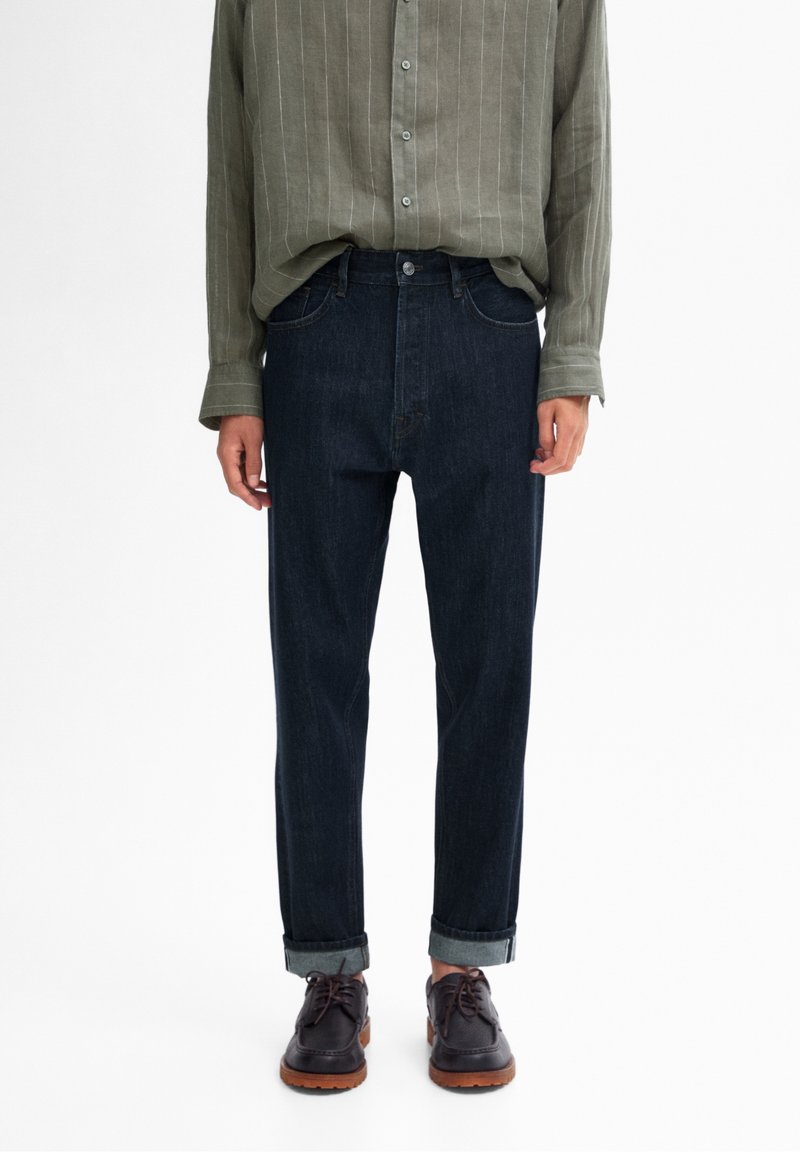 Personne portant un jean bleu foncé retroussé, une chemise kaki boutonnée et des chaussures à lacets noires avec des semelles marron, debout devant un fond blanc.