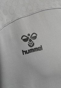 Grijs sportkledingstof heeft een textuurdesign, met een subtiel gestippeld patroon en een zwart Hummel-logo in de rechterbenedenhoek.