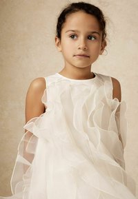 Biely priesvitný šaty s vrstvenými volánmi a jemnými okrajmi. Pevné ramienka, ľahká tkanina a mäkká textúra vytvárajú plynulý vzhľad.