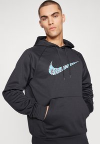 Juodas Nike džemperis su dideliu turkio spalvos raštuotu logotipu. Yra priekinė kišenė ir raištelio gobtuvas. Lygus, lengvas audinys.