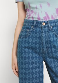 Osoba ubrana w niebieskie dżinsy w diamentowy wzór z miedzianym guzikiem oraz jasnoniebieską koszulę w tie-dye z purpurowymi plamami.