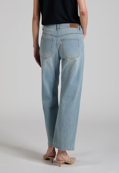 Persona che indossa jeans azzurri a gamba dritta e scarpe slingback beige con tacco, in piedi con le mani in tasca, rivolta di spalle.