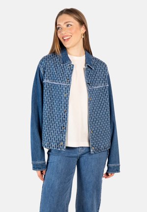 Smilende kvinne iført blåmønstret denimjakke over hvit skjorte med blå jeans, stående mot en ensfarget lys bakgrunn.