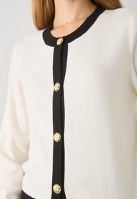 Cardigan en tricot crème avec bord noir, présentant un col rond et six boutons dorés à l'avant. Texture douce, design ajusté.