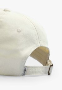 Cream-colored baseball cap made of textured fabric, featuring a curved brim, adjustable strap, and a small logo tag on the side.