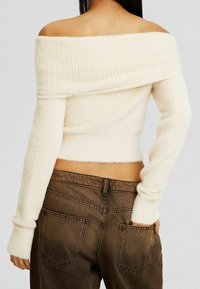 Kvinna iklädd en off-shoulder vit fluffig tröja och bruna högmidjade jeans, med vänster hand i bakfickan, sedd bakifrån.