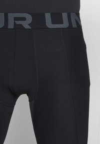 Svarta sportleggings med ett grått elastiskt midjeband med texten "UNDER", slät yta och åtsittande design. Sömmar är synliga.