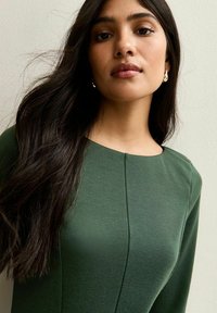 Zielona sukienka z długim rękawem i okrągłym dekoltem, wykonana z fakturowanej tkaniny. Gładka powierzchnia z pionowymi szwami, połączona ze złotymi kolczykami.