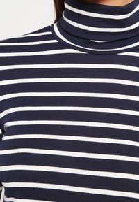 Maglione a collo alto di colore blu navy con righe bianche orizzontali. Tessuto liscio e elastico con un design aderente, caratterizzato da un colletto alto.