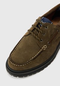 Brauner Bootsschuh aus Wildleder mit Schnürdesign, Metallösen und einem kontrastierenden, weiß abgesteppten Rand. Robuste schwarze Gummisohle.