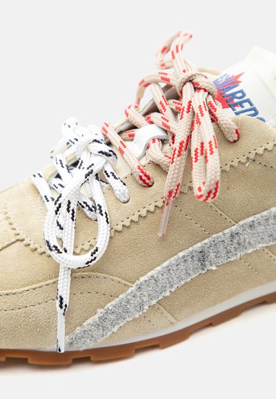 Μπεζ δερμάτινο αθλητικό παπούτσι με δύο κορδόνια διαφορετικού χρώματος—άσπρα με μαύρες ρίγες και μπεζ με κόκκινες ρίγες—και υφασμάτινη γκρι λεπτομέρεια στο πλάι.