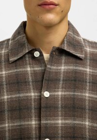 Tuva tuvplāns, kurā redzams cilvēks ar brūna svītrojuma pogām kreklu, kurā ir redzamas baltas pogas un bēša apakškrekls, kas redzams pie apkakles.