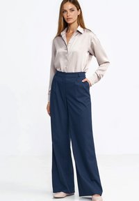 Jasnoróżowa satynowa bluzka z guzikami z przodu, zestawiona z szerokimi spodniami w granatowe paski. Spodnie mają wysoki stan i boczne kieszenie.