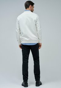 Sudadera blanca con puños acanalados, llevada sobre una camisa de cuello azul claro, combinada con jeans oscuros y calzado negro, vista desde la parte posterior.
