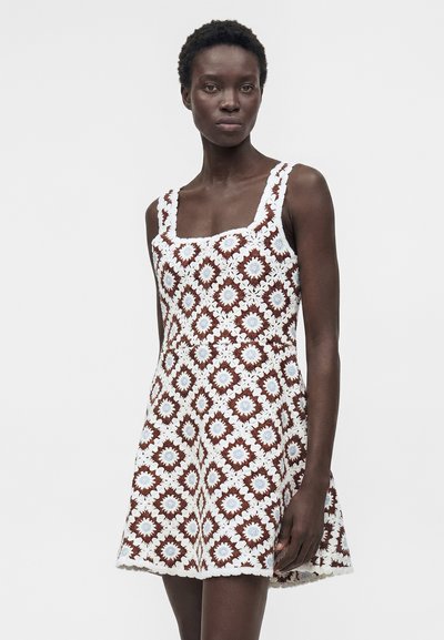 Μαύρη γυναίκα φοράει αμάνικο πλεκτό φόρεμα με καφέ και λευκό γεωμετρικό floral μοτίβο, στέκεται μπροστά σε απλό ανοιχτό φόντο.