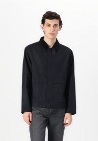 Black lightweight jacket with a collar, front snap closure, two large pockets, and long sleeves, worn over gray jeans.