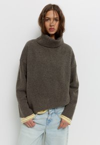 Brauner Strickrollkragenpullover mit gerippten Bündchen und Saum, der eine lockere Passform und eine weiche Textur aufweist, über einem gelb gestreiften Hemd getragen.