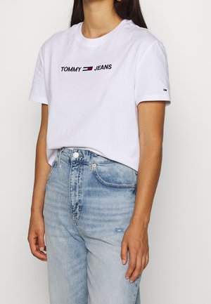 Vrouw draagt een wit Tommy Jeans T-shirt met logo op de borst en lichtblauwe high-waist jeans, staand tegen een effen achtergrond.