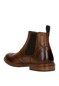 Stivale Chelsea in pelle marrone con pannelli laterali elastici, dettagli brogue e tacco in legno impilato. Texture liscia con accenti neri a contrasto.