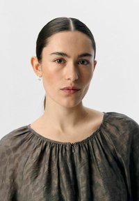 Blouse texturée grise avec un col froncé et un petit détails de fermeture éclair à l'avant, portée par un modèle sur un fond neutre.