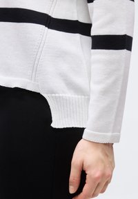 Λευκή ζακέτα με μαύρες οριζόντιες ρίγες στα μανίκια και το στήθος, με ριμπ τελείωμα, φορεμένη με μαύρο παντελόνι, με το χέρι σε χαλαρή θέση δίπλα στο σώμα.