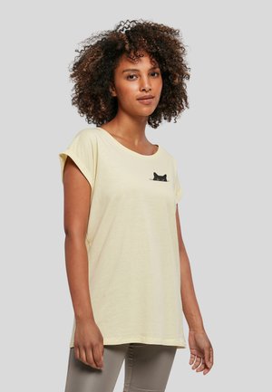 Kvinde med krøllet hår iført lysegul t-shirt med lille sort kattegrafik på brystet og lysegrå bukser, står mod grå baggrund.