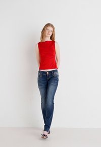 Rdeča brezrokavna majica, prilegajoča se zasnova, v kombinaciji z modrim jeansom. Jeans vsebujejo žepe in blag odtenek. Model stoji na svetli podlagi.