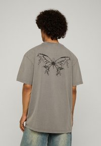 Cilvēks ar kārpainiem matiem valkā pelēku pārāk lielu T-kreklu ar melnu abstraktu tauriņu grafiku uz muguras un izbalējušām zilām džinsiem.