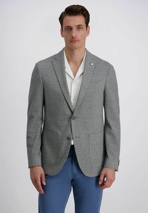 Man draagt een grijs getextureerd colbert, een wit overhemd met open kraag en blauwe broek, staand tegen een effen lichte achtergrond.