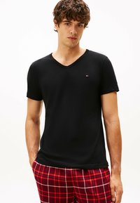 Camiseta negra de manga corta con escote en V, fabricada en algodón suave, estilo ajustado, pequeño logo en el pecho, combinada con pantalones de pijama de cuadros rojos.