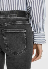 Personne portant un jean denim noir avec une poche arrière et une chemise rayée à manches longues, main détendue sur le côté avec des bagues en or visibles.