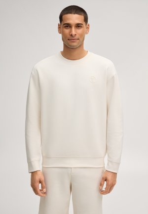 Mann trägt einen cremefarbenen Pullover und passende Hose, steht vor einem schlichten hellen Hintergrund.