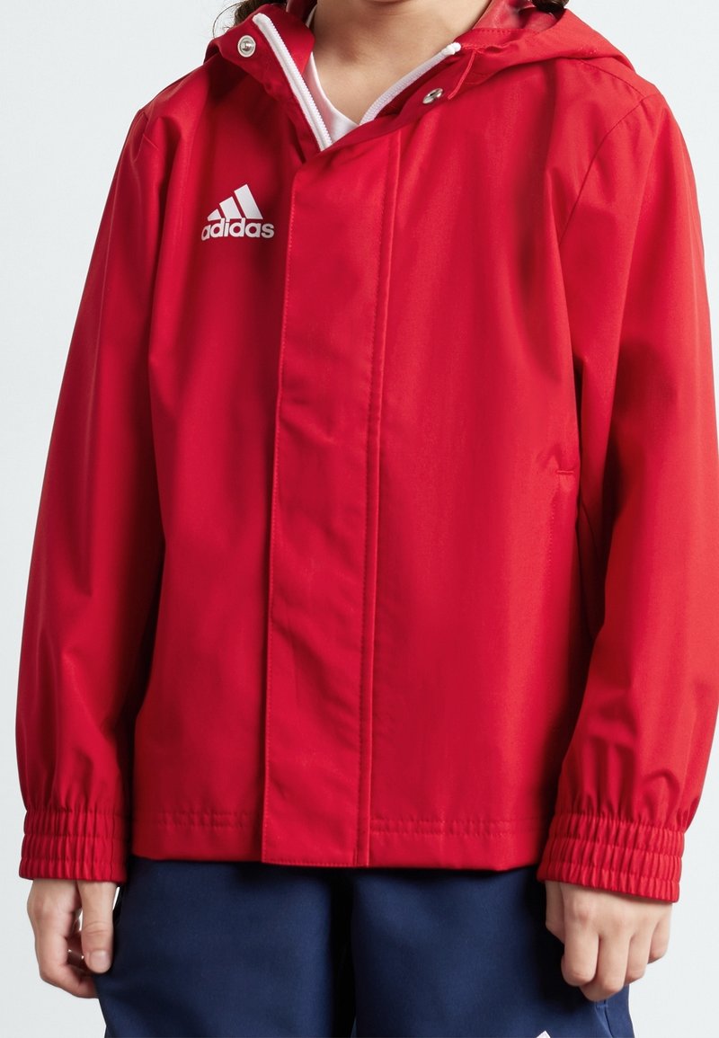 Rode waterdichte jas met een capuchon, elastische manchetten en een wit adidas-logo op de linkerborst. Heeft een gladde, zachte textuur.
